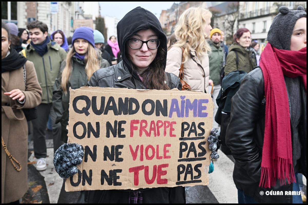 dans la manifestation du 25 novembre, dans la foule, se tient une femme qui regarde l'appareil photo, elle tient une pancarte "Quand on aime, on ne frappe pas, on ne viole pas, on ne tue pas"